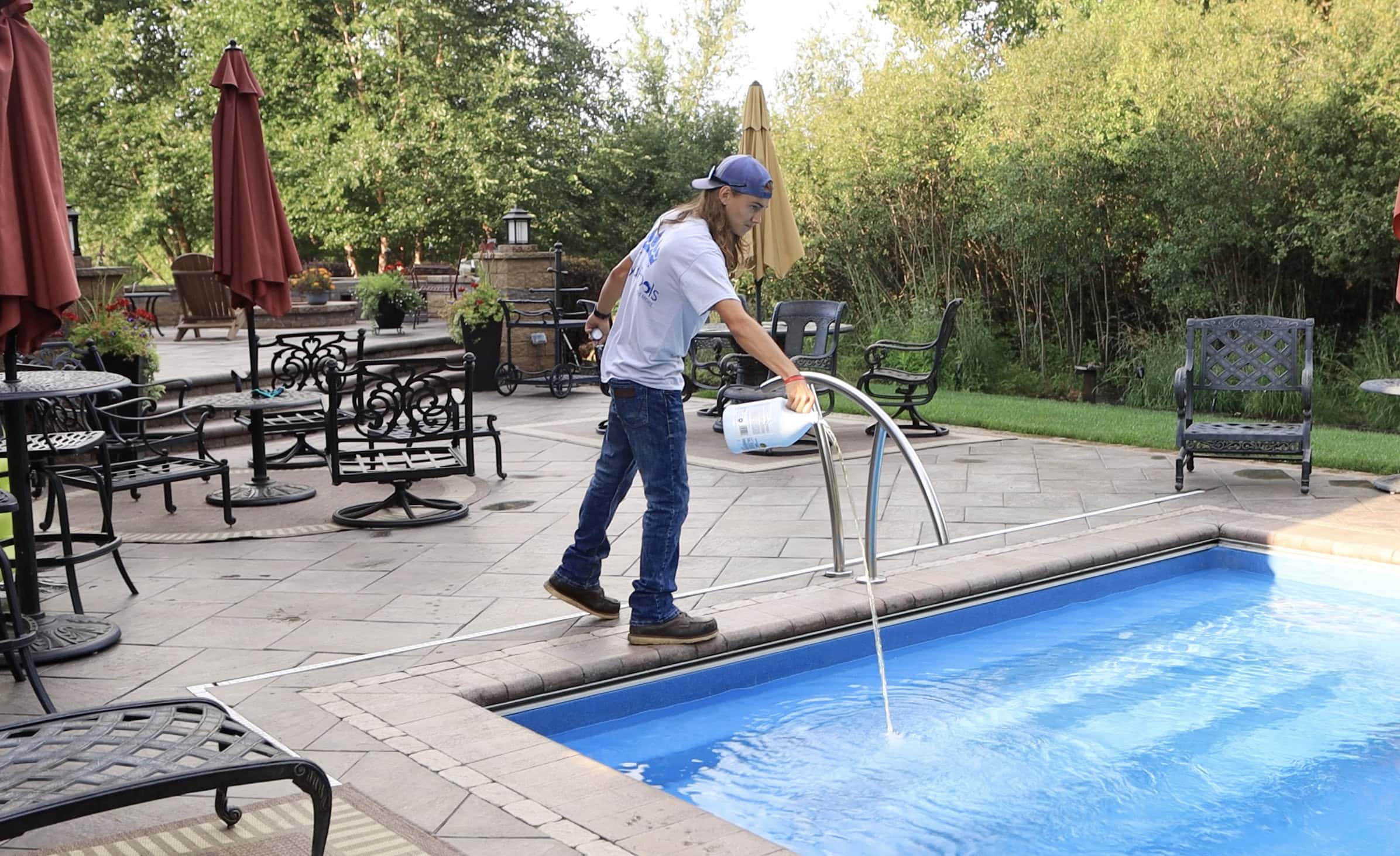Photo of a man standing over the shallow end of a light-blue inground pool pouring a yellow liquid into the pool. Behind the pool is a stamped concrete patio with chairs and a retaining wall. Trees can be seen behind the patio.